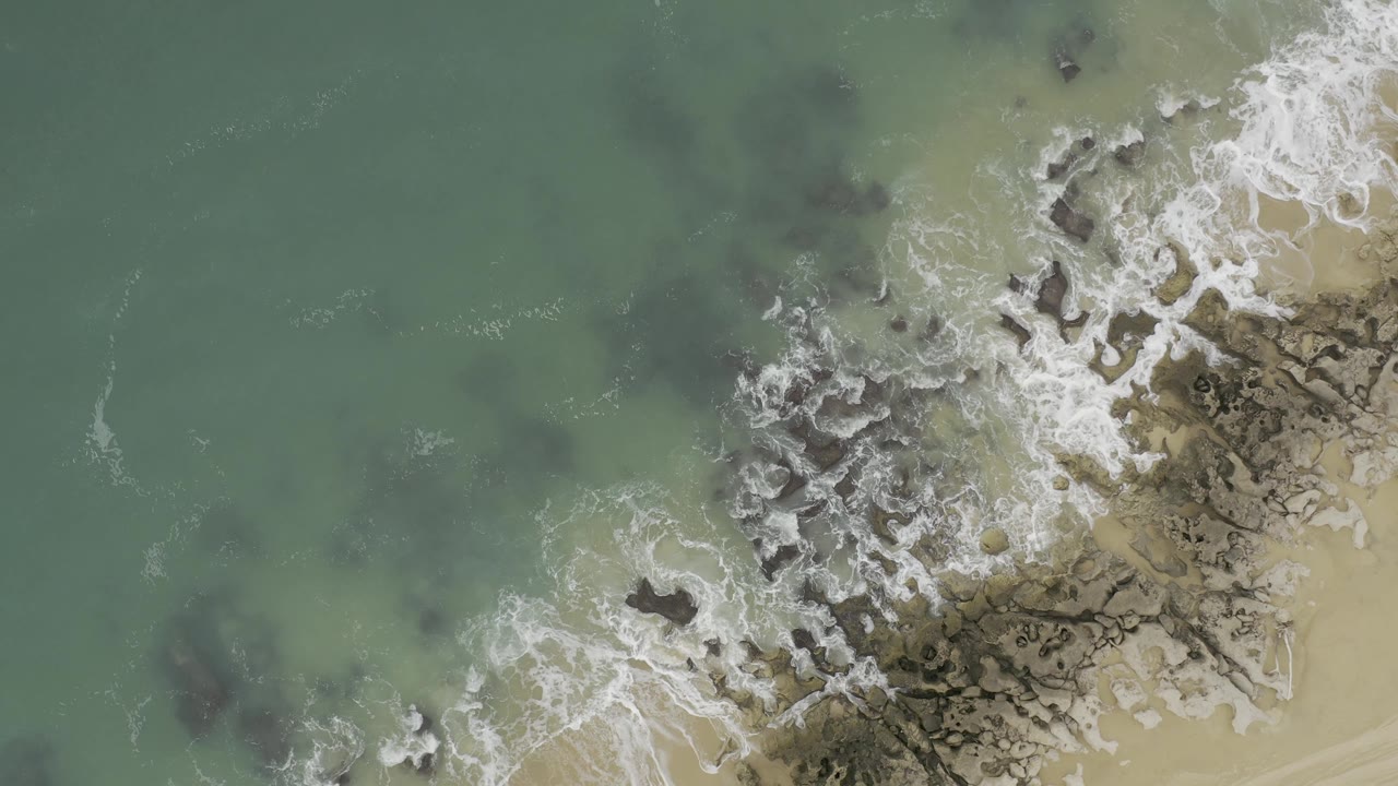 Top-down Baja California Sur Mexico shoreline frame with white surf curling over gray rocks and pale tan beach sand along the right side, no visible people.