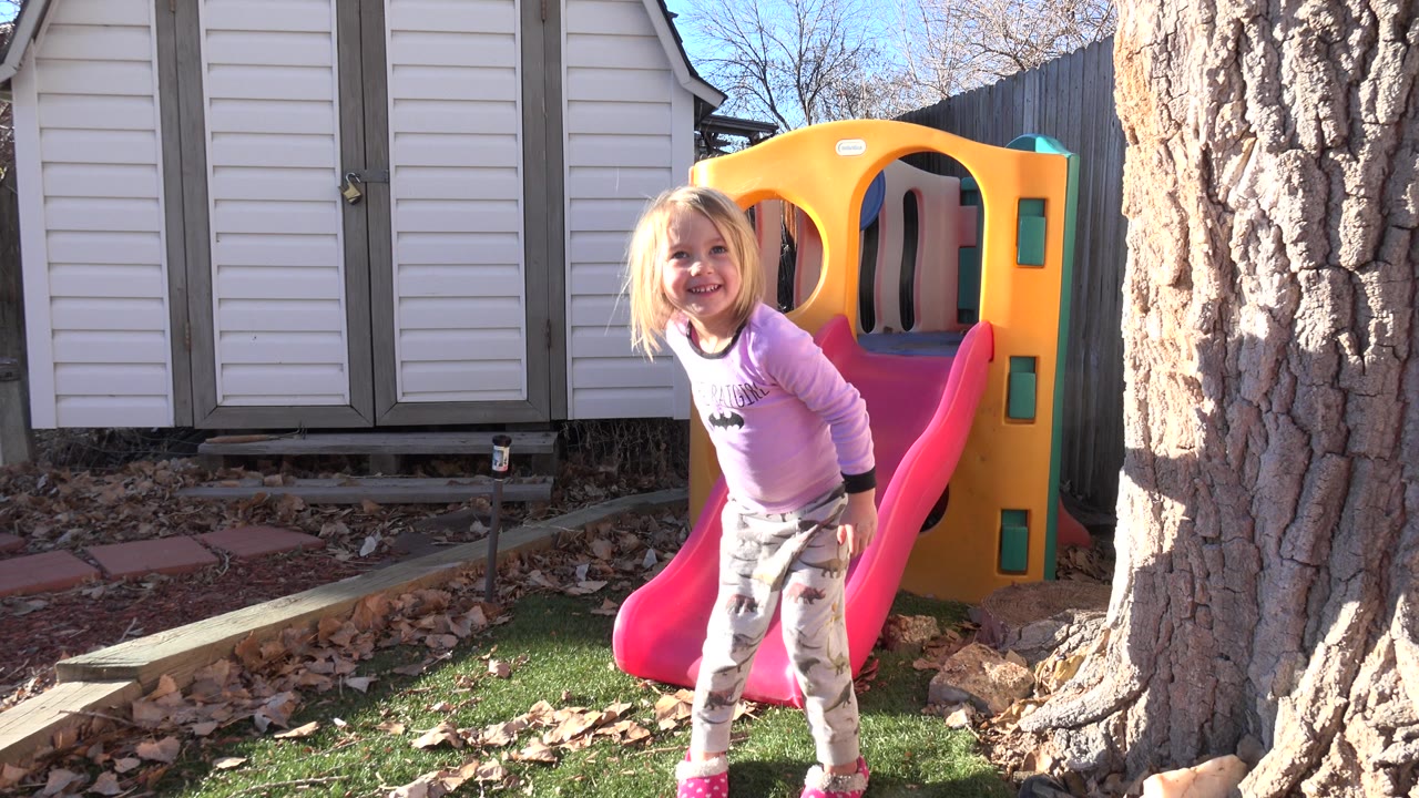 The girl leans forward smiling beside the pink slide, with the yellow playhouse structure and tree trunk still prominent in the backyard frame.