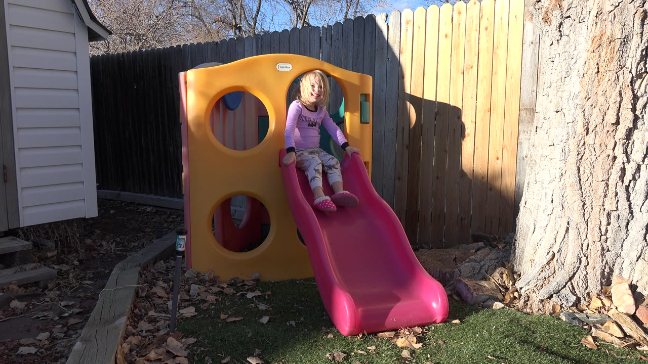 At 0:00 in Westminster, Colorado, a young blonde girl sits at the top of a pink slide on a yellow plastic backyard play structure beside a wooden fence and tree.