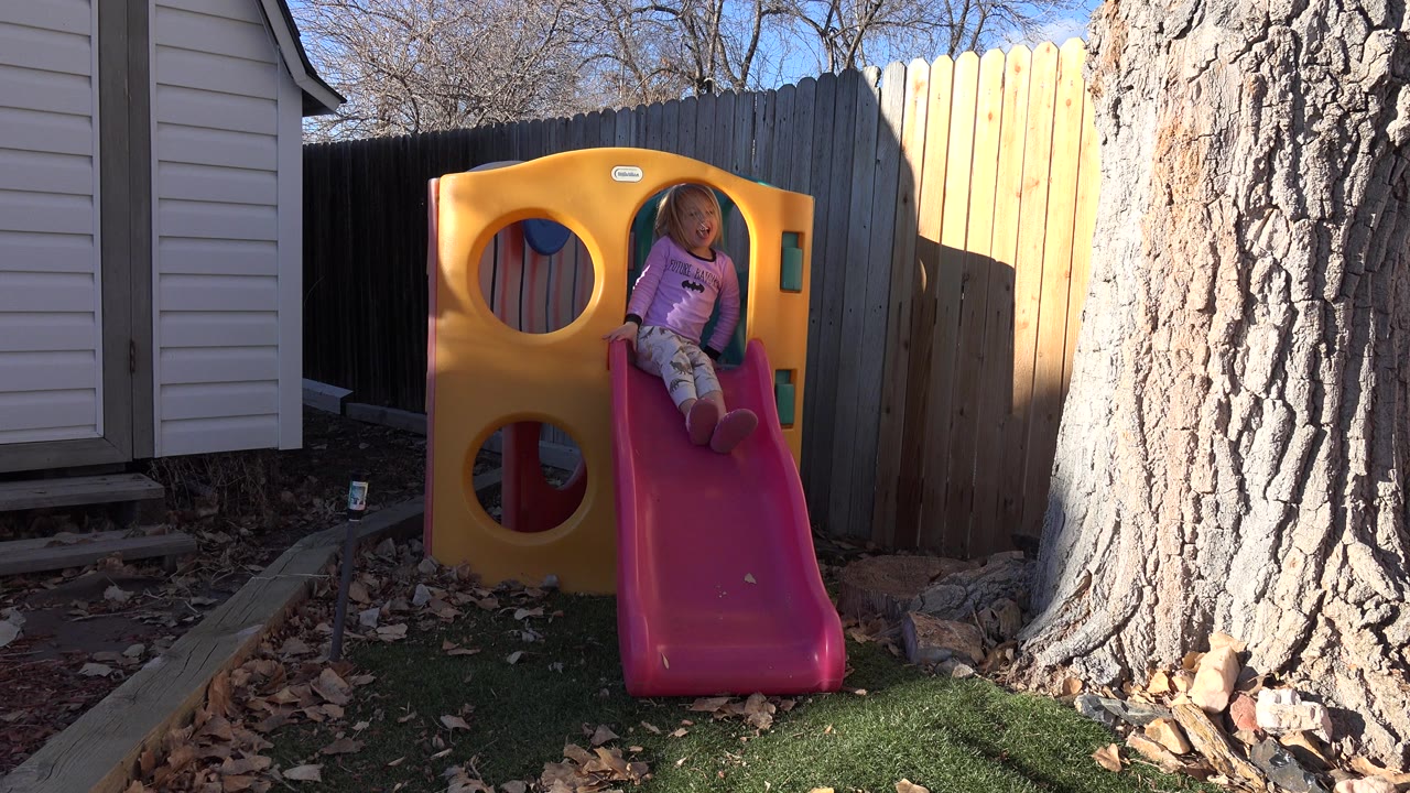 Child sitting near the top of a pink slide in a backyard.