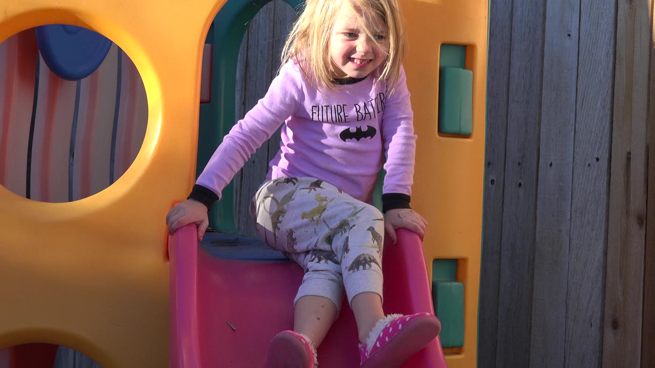 The girl smiles while seated on the red slide, with one foot extended and the yellow plastic play structure filling the left side of the frame.