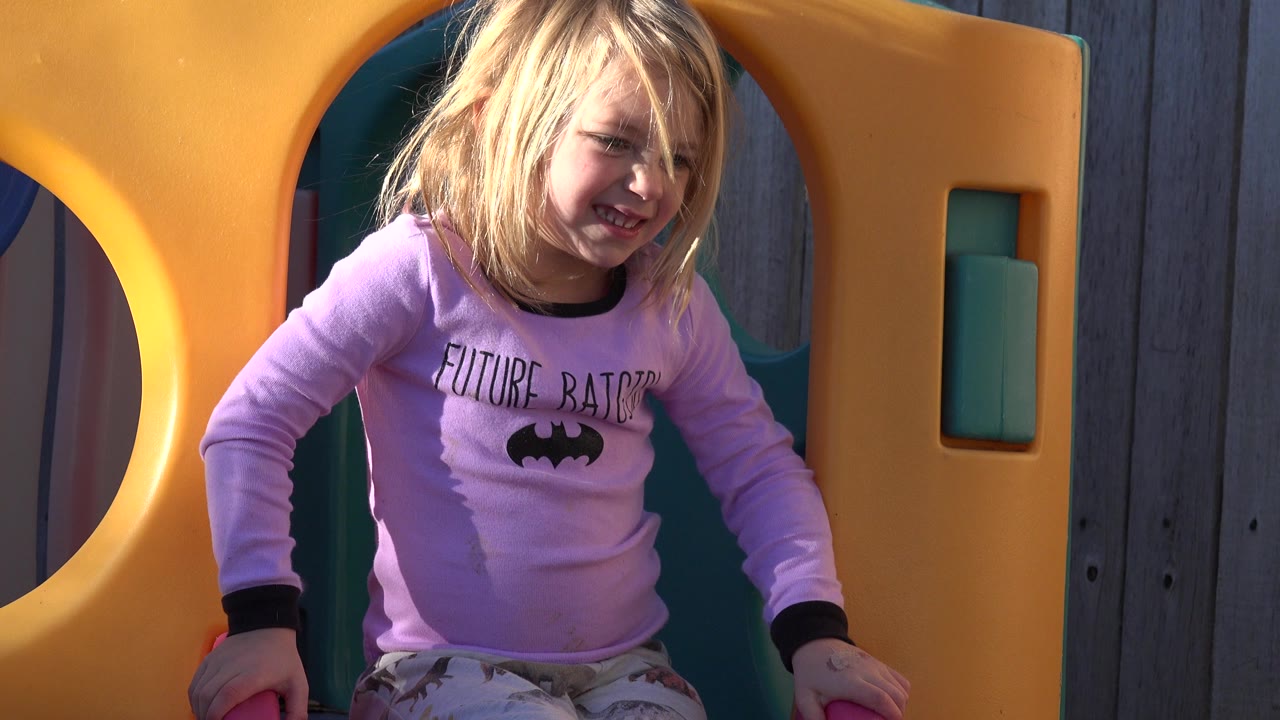 The young girl smiles while leaning forward in the yellow and green jungle gym, with the wood fence still visible behind the play structure in Westminster, Colorado.