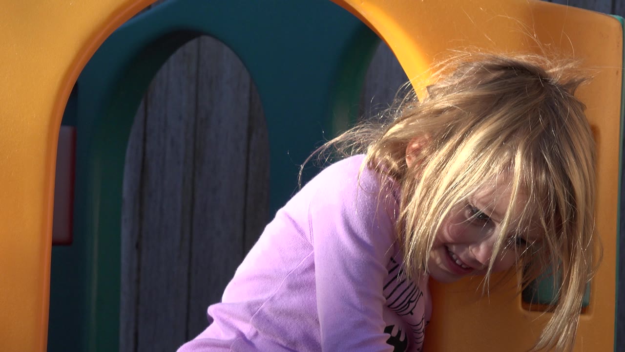 One second in, the girl remains close to the yellow play structure opening, with her face partly visible under loose blond hair in the Westminster backyard scene.