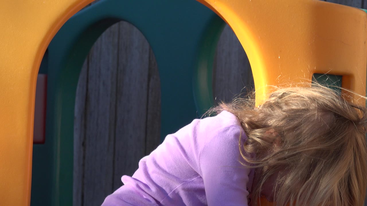 At 0:01, the girl's hair falls across her face as she leans forward beside the yellow playground arch in Westminster, Colorado.