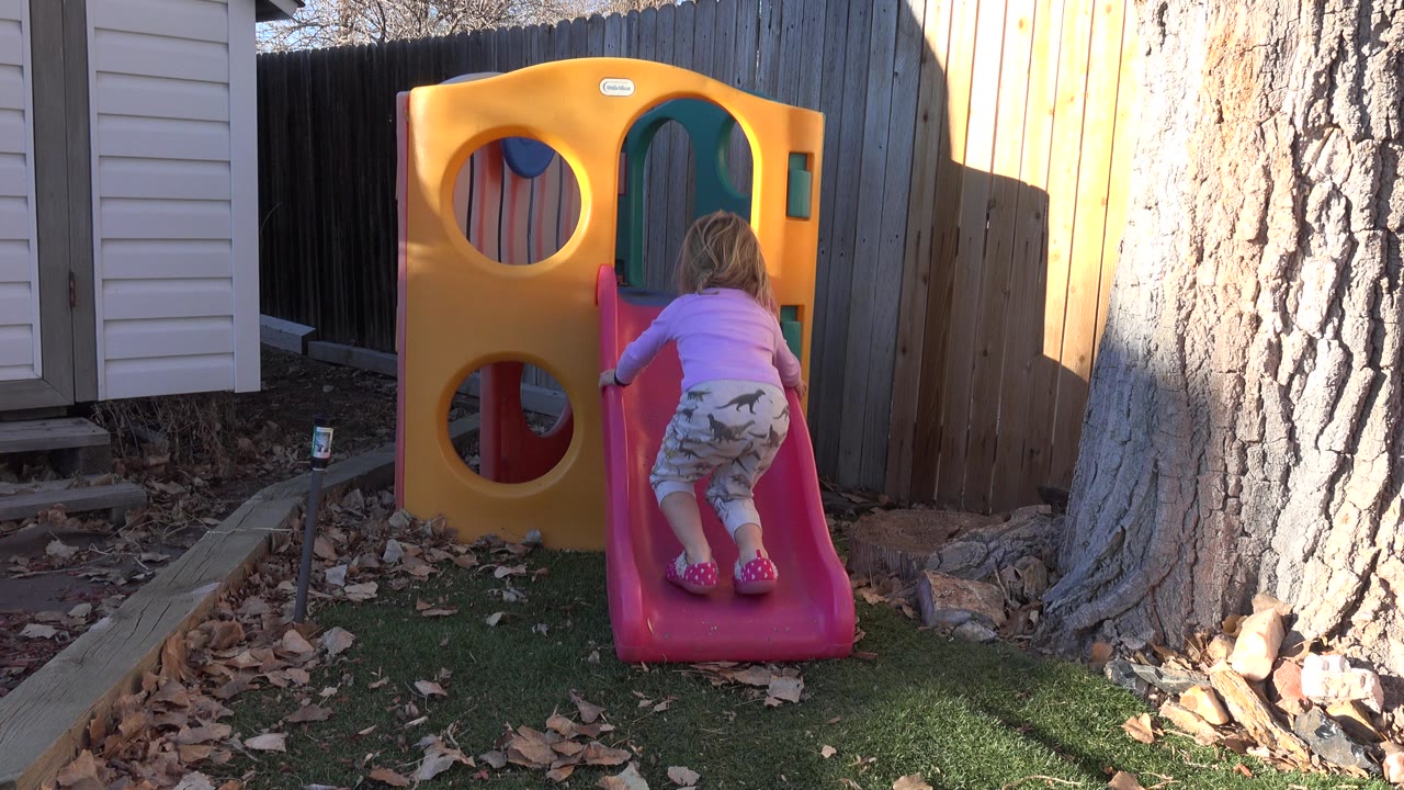 Young girl climbing a pink backyard slide on a yellow playhouse.