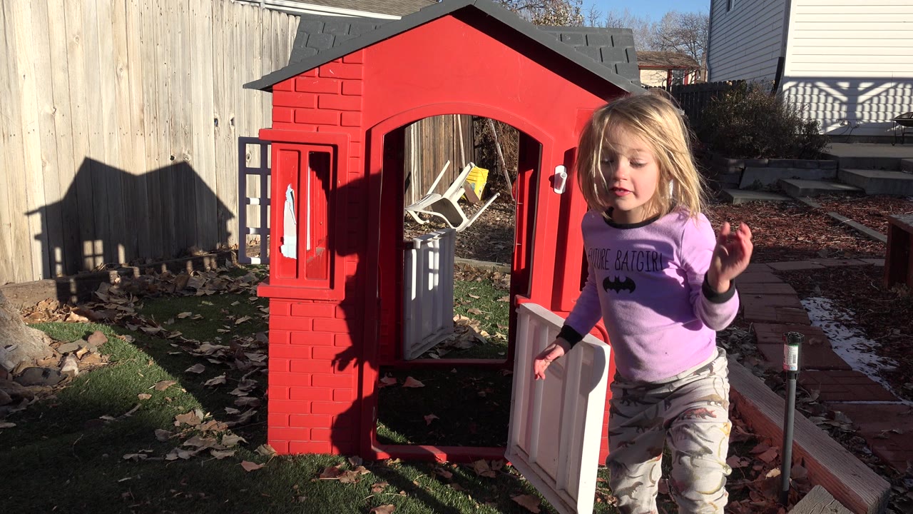 Child standing beside a red backyard playhouse in bright sun.