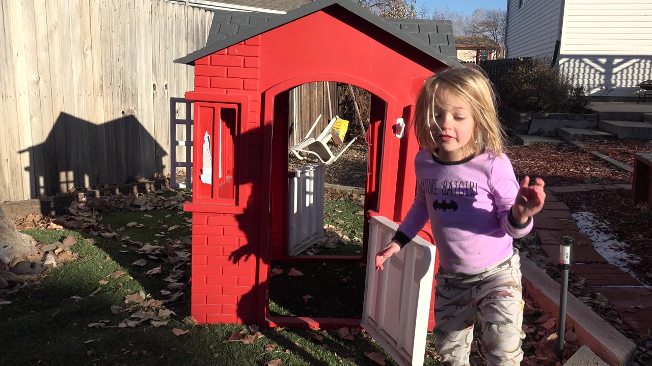 The child remains in the right foreground beside the red playhouse, with the open doorway, fence, leaves, and low sunlight visible in the Westminster backyard.