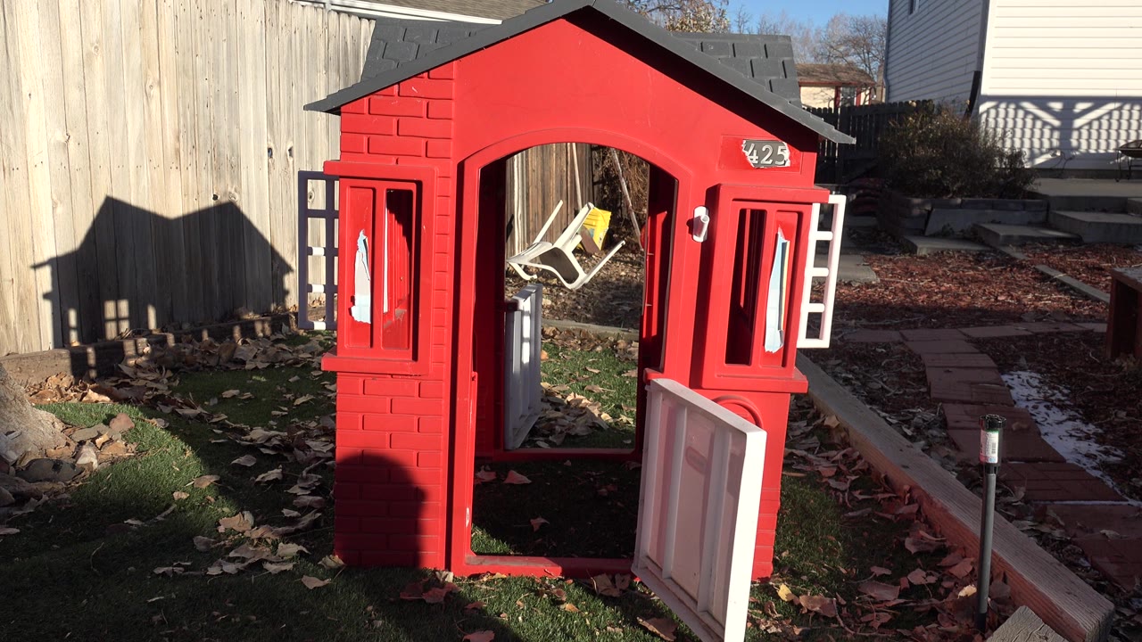 Red plastic playhouse in a Westminster, Colorado backyard with the white swinging door open, two windows visible, fallen leaves on the grass, and no clearly visible people.