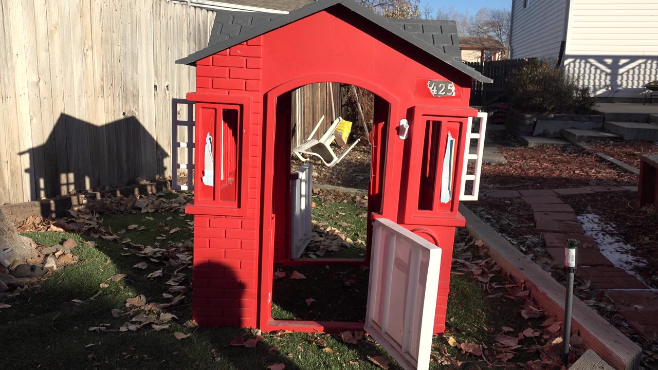 Red playhouse remains centered in the Westminster backyard, with a dark roof, open white door, bright fence light, long shadows, and no clearly visible people.