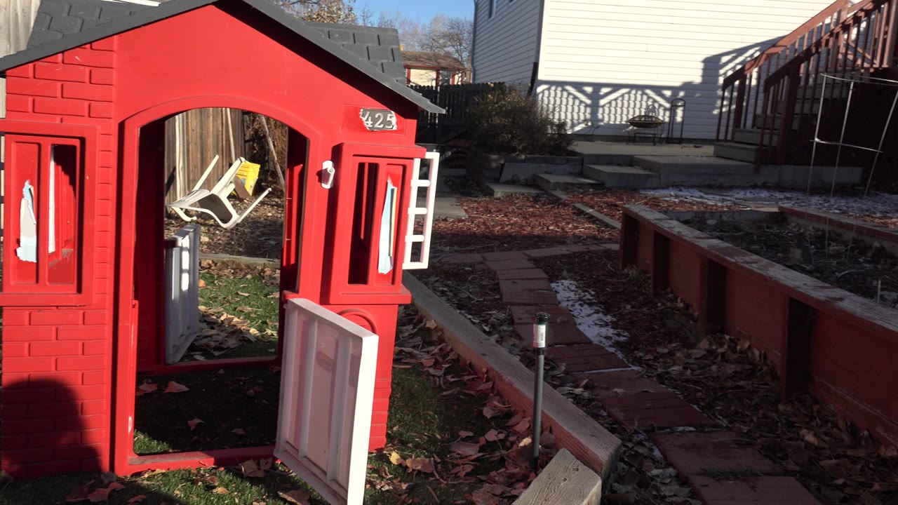 A Westminster, Colorado backyard view holds on the red playhouse at left, raised garden beds in the center, and a house deck and fence in the background with no visible people.