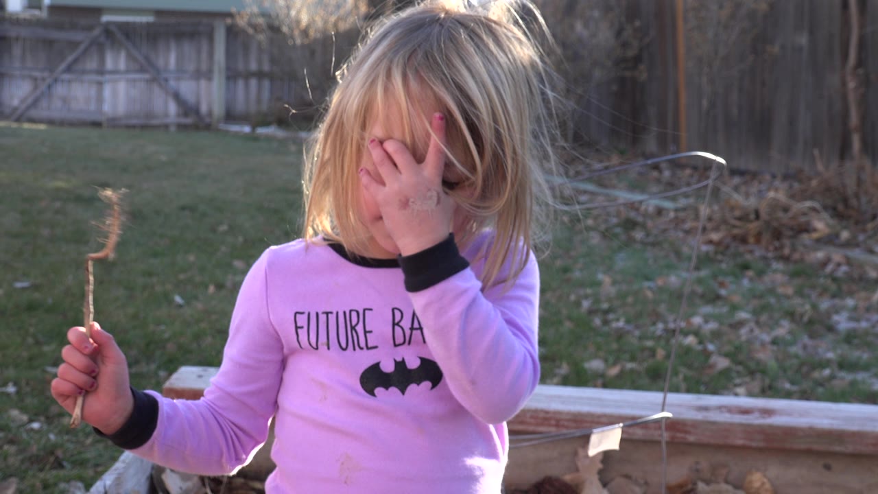 Girl holding up a stick with one hand over her face in a backyard.