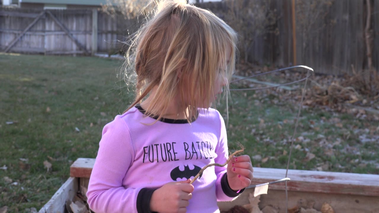 Blond child in a pink shirt looks left while holding a stick outdoors.