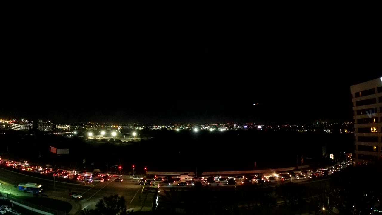 Opening frame of Los Angeles at night with red taillights along the lower road, distant city lights across the horizon, a white building on the right, and no visible people.