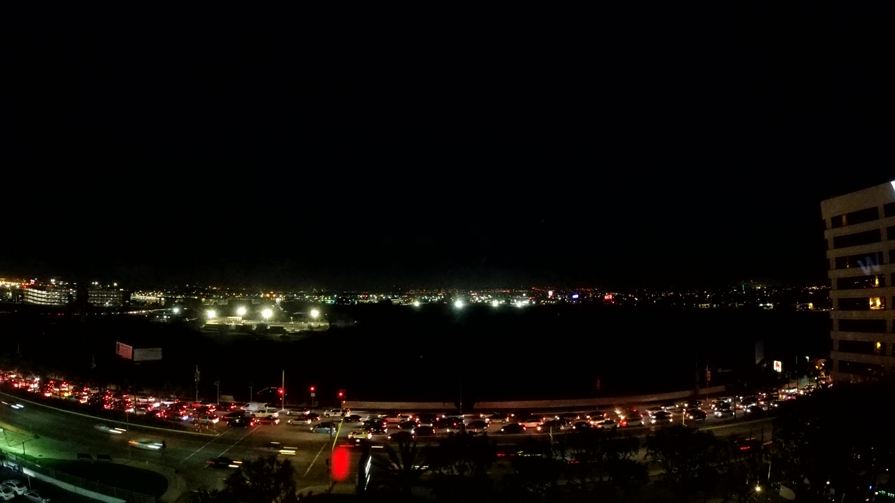 A wide Los Angeles evening view shows cars packed along the lower road, traffic signals glowing red and green, distant city lights, and no visible people.