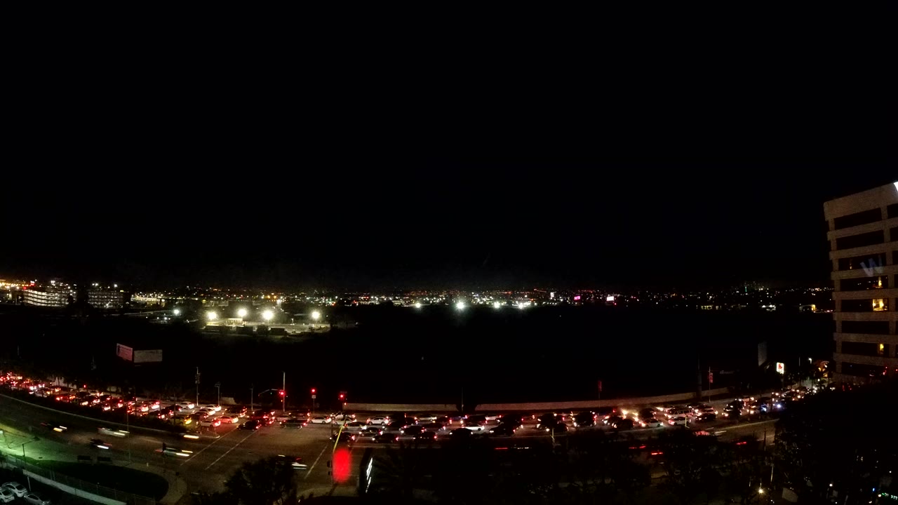 Elevated Los Angeles night view with a busy road across the lower frame, red brake lights, white headlights, distant city lights, a dark sky, and no clearly identifiable people.