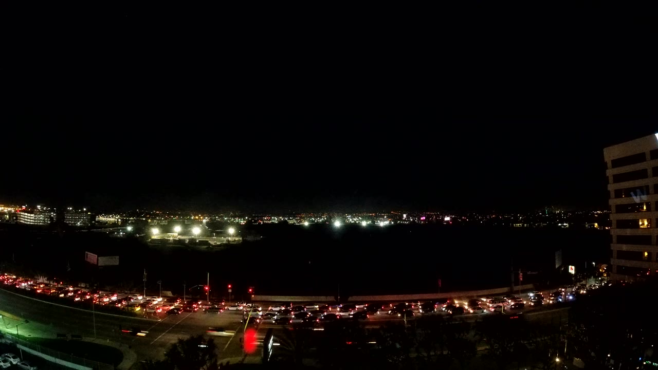 Los Angeles road traffic fills the lower frame at night, with red lights clustered near an intersection, bright distant lights on the horizon, and a cropped building on the right edge.