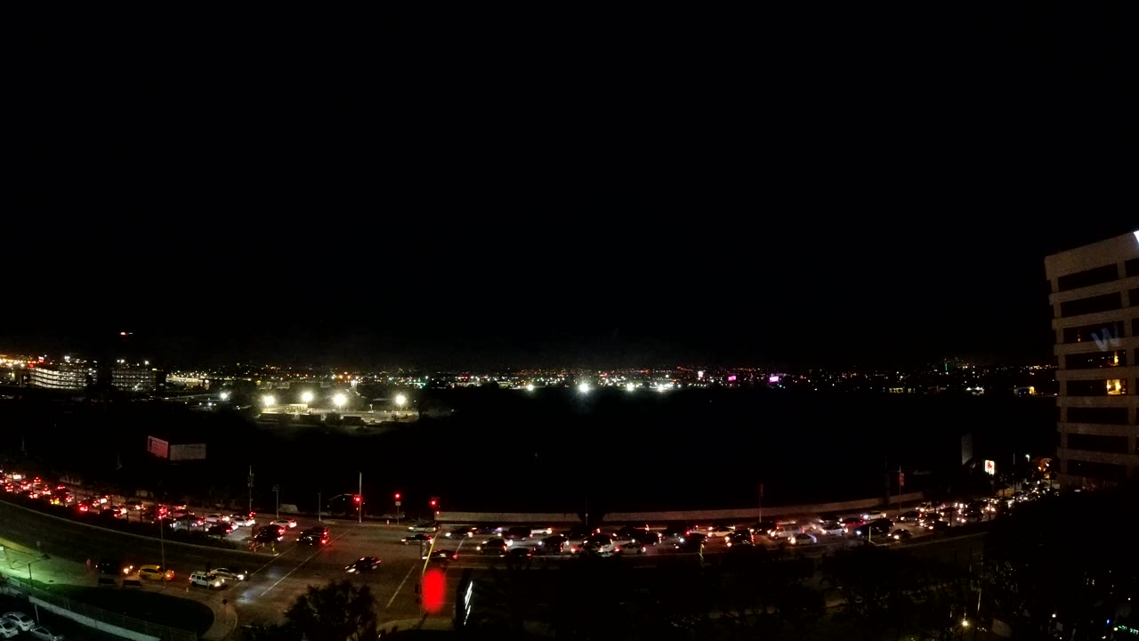 A Los Angeles nighttime intersection sits in the foreground with rows of car lights, a red signal glow near the lower center, distant horizon lights, and the right-side building partly visible.