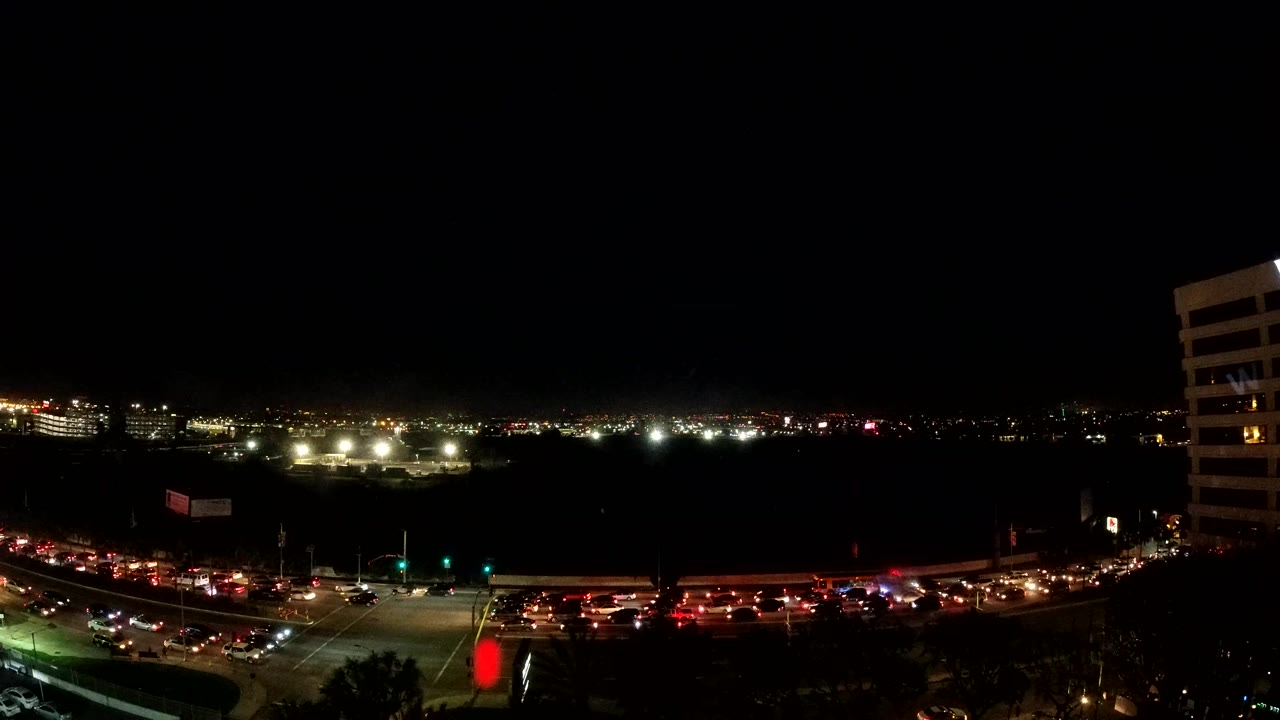 Los Angeles evening road scene with dense traffic, red signals near the center, distant lights across the horizon, and the white building still visible on the right, with no visible people.