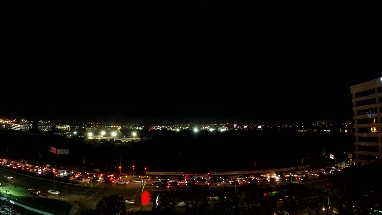 Los Angeles traffic passes through the lower frame under a black sky, with greenish road lighting at left, a band of distant lights, and a white high-rise at right, with no visible people.