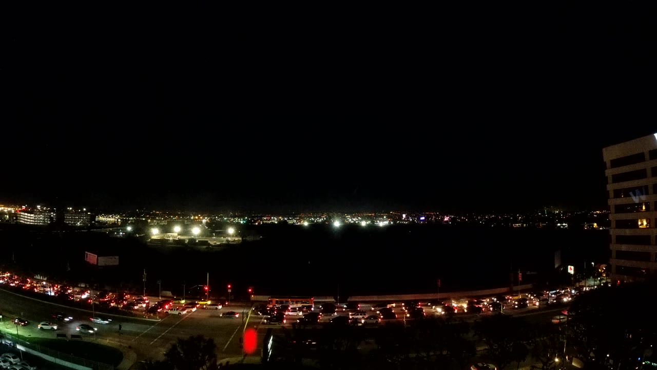 Los Angeles night traffic curves through the foreground with clusters of red brake lights, bright distant lights on the left, and the white high-rise cropped along the right side, with no visible people.