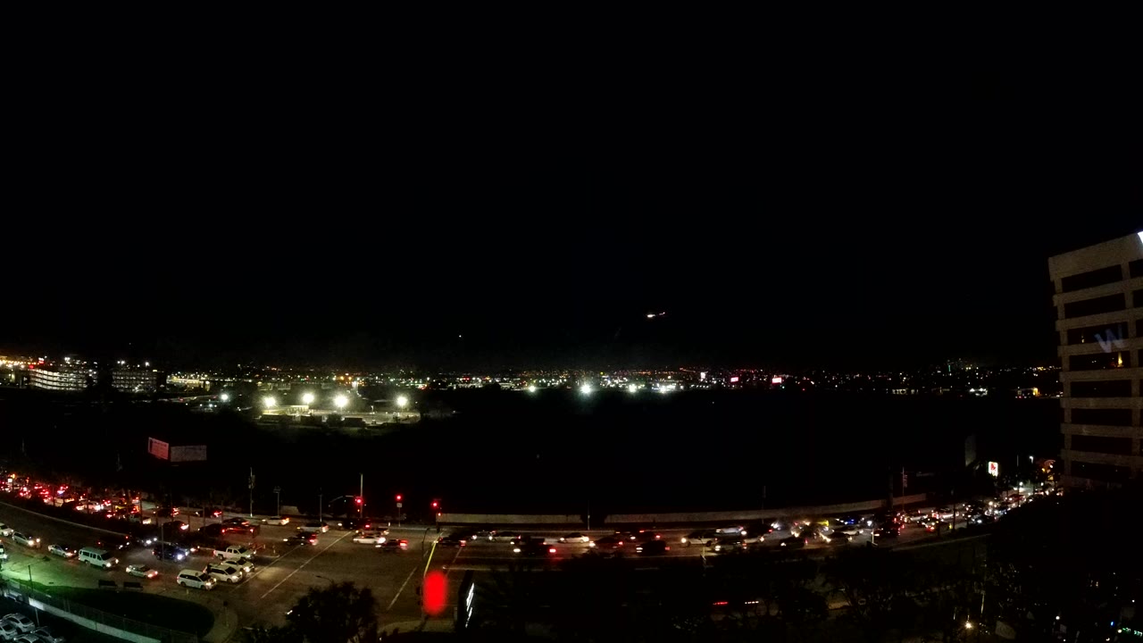 Los Angeles night traffic seen from an elevated view, with red and white vehicle light streaks crossing a road below distant city lights and no visible people.