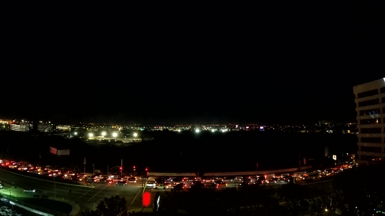 Elevated Los Angeles road view at night, showing layered red traffic streaks across the lower frame, bright horizon lights, and a tall building edge on the right with no visible people.