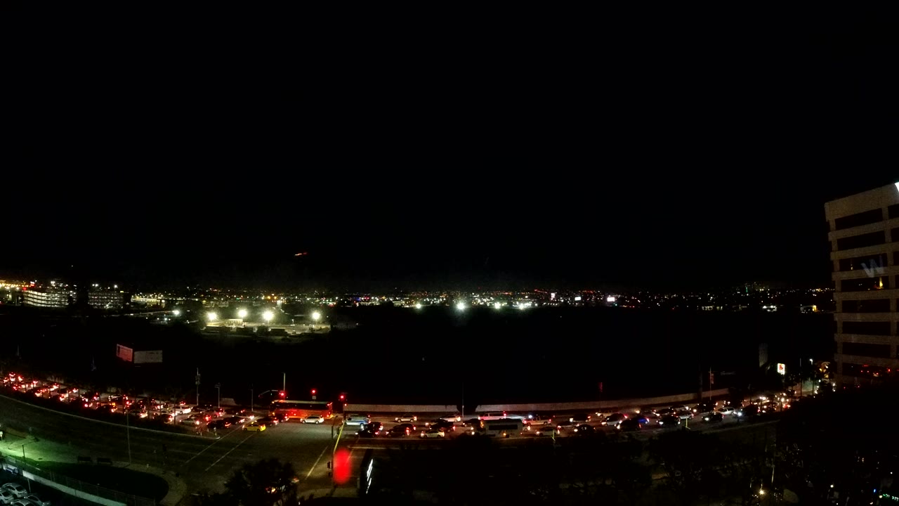 Los Angeles night traffic runs along a waterfront road under a black sky, with red tail lights, white headlights, distant city lights, and no visible people.