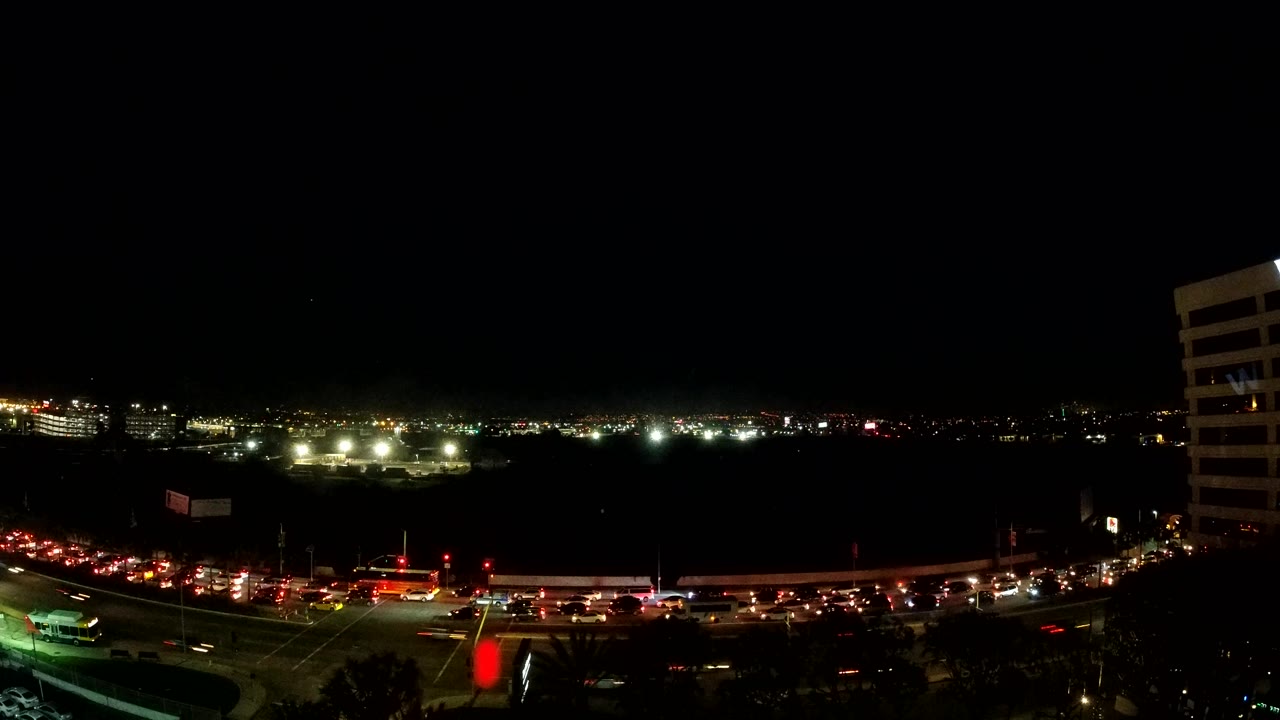 Los Angeles traffic queues through a lit intersection at night, with red signal lights, headlight streaks, a dark waterfront midground, distant lights, and no visible people.