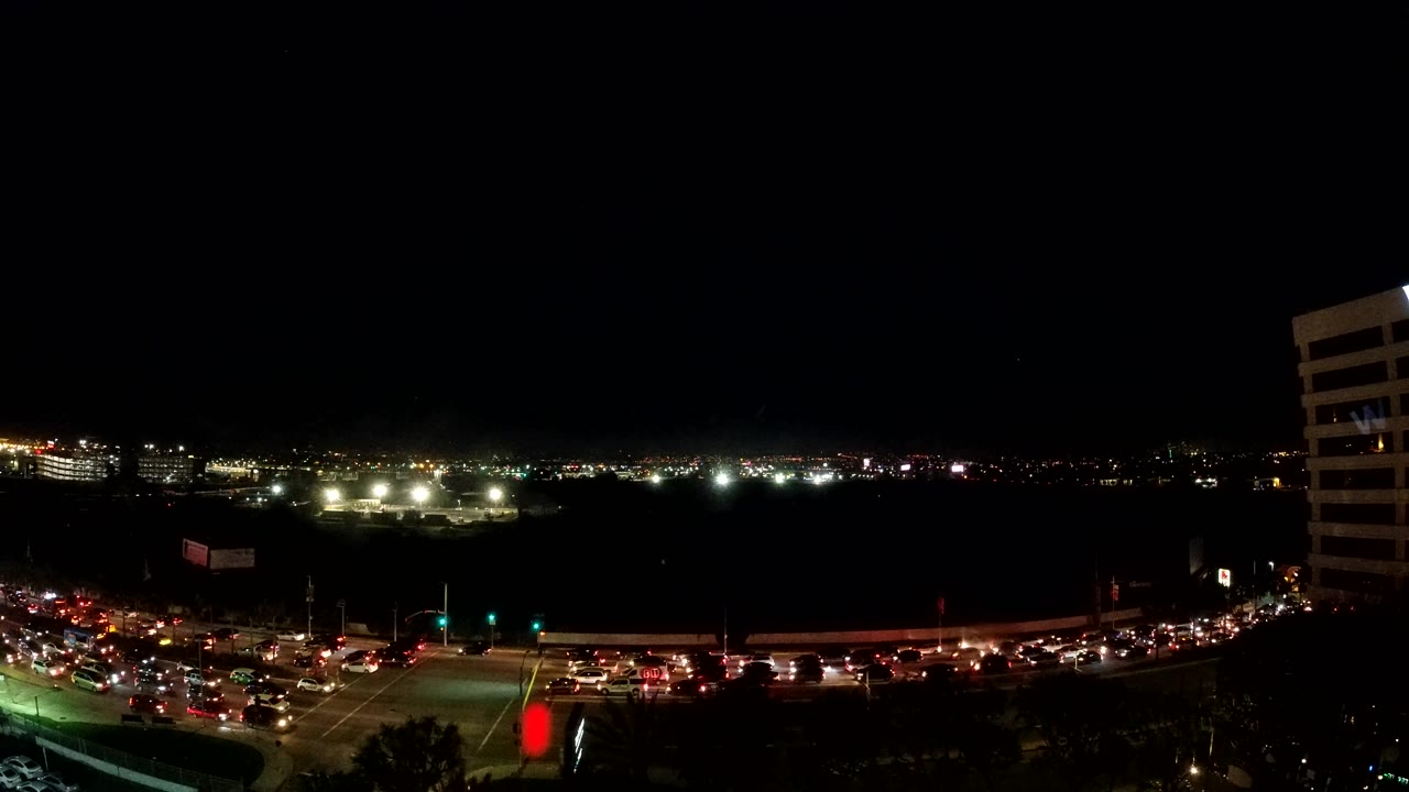 Elevated Los Angeles view with green traffic signals, cars spreading across the avenue, a dark midground, and distant lights across the horizon, no visible people.