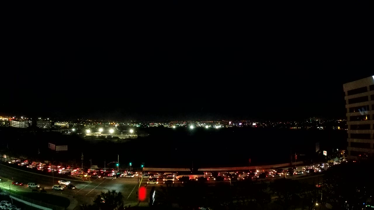 Opening frame of a wide elevated Los Angeles night traffic view with red taillights, white headlights, distant city lights, a dark sky, and no visible people.
