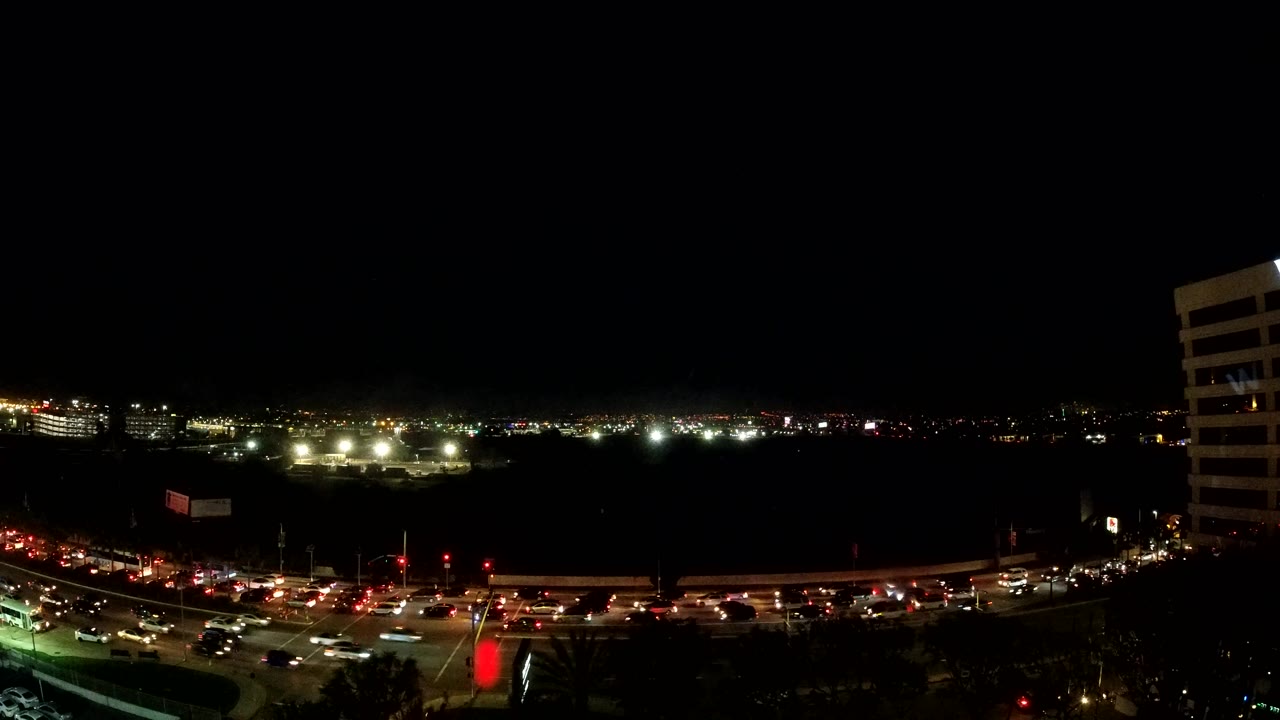 Wide elevated Los Angeles traffic frame with rows of cars, bright road lighting, a tall building cropped at the right edge, and no visible people.
