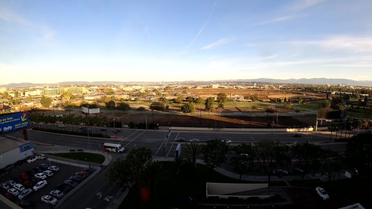 Elevated Los Angeles view at sunset with multilane roads, parked cars in the foreground, low buildings in the distance, and no visible people.