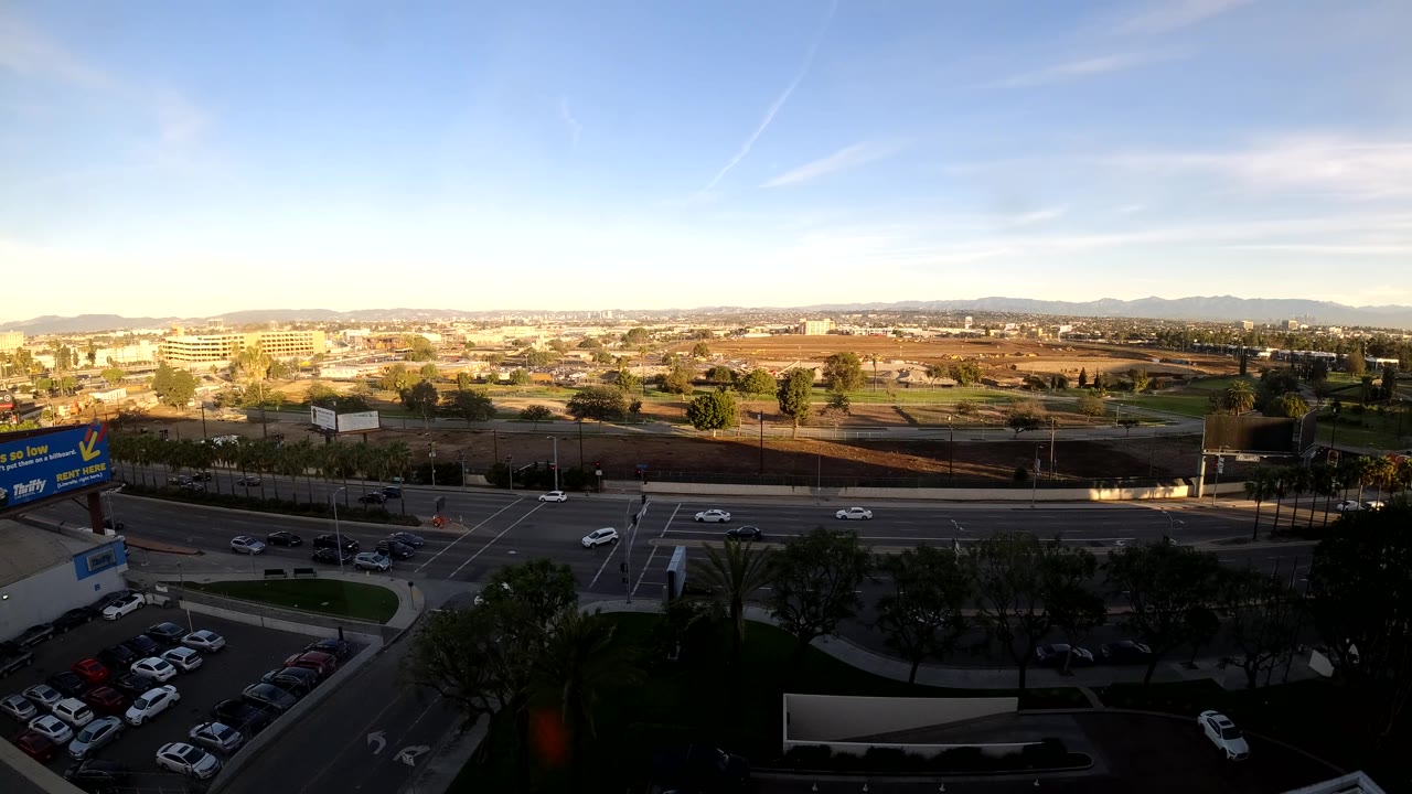 Elevated Los Angeles frame with vehicles moving through the road grid, a commercial sign at left, parking below, distant hills, and no visible people.