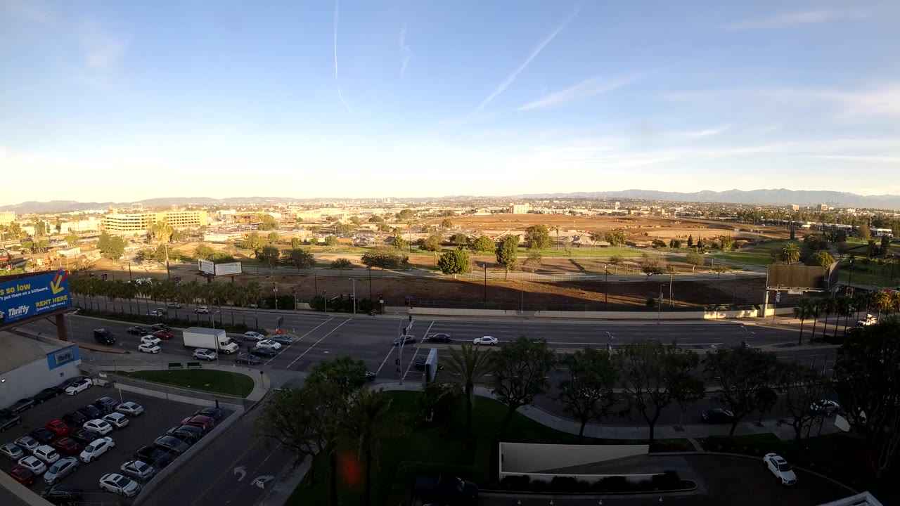Los Angeles traffic frame with a white truck near the intersection, several cars on the roadway, shadowed trees and parking in the foreground, low-rise buildings across the road, and distant hills under blue sky.