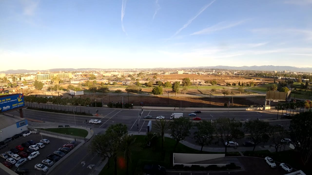 Opening frame of Los Angeles traffic from an elevated wide angle, with cars and trucks crossing a large intersection, a parking lot in the foreground, distant hills, warm low light, and no visible people.