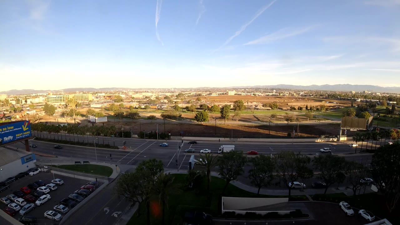 Wide elevated view of traffic lanes, parking lot, trees, and distant city edge under blue sky.