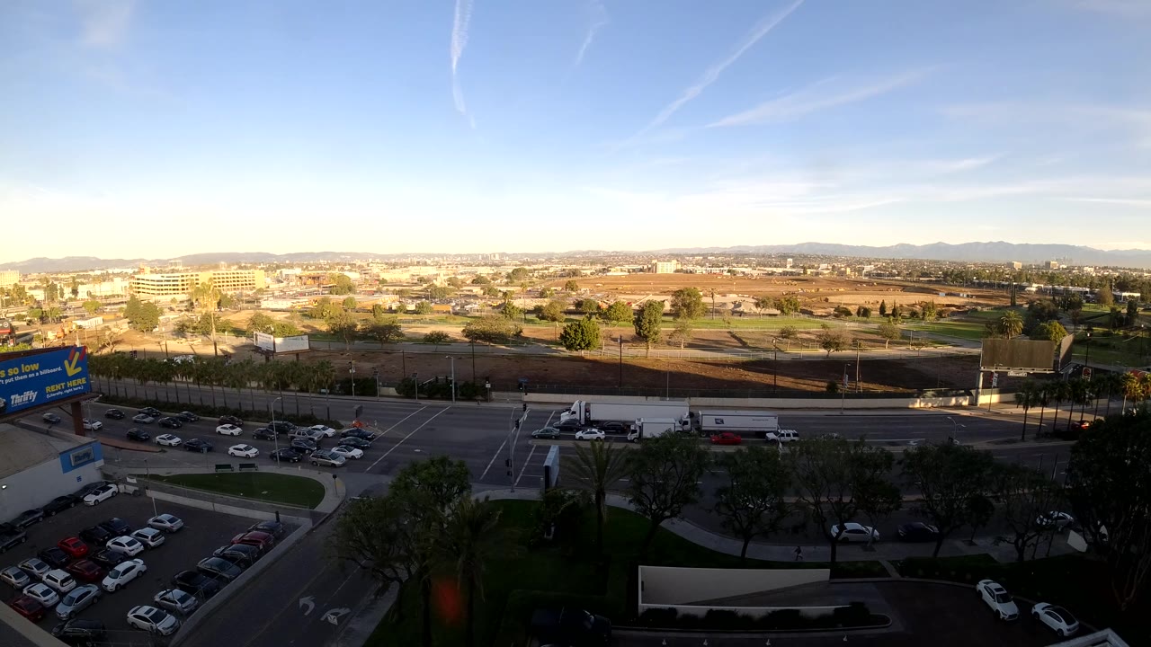 Third frame of the elevated Los Angeles intersection, with cars crossing the lower road, trucks in the midground, shadowed trees and parked cars below, bright sky above, and no visible people.