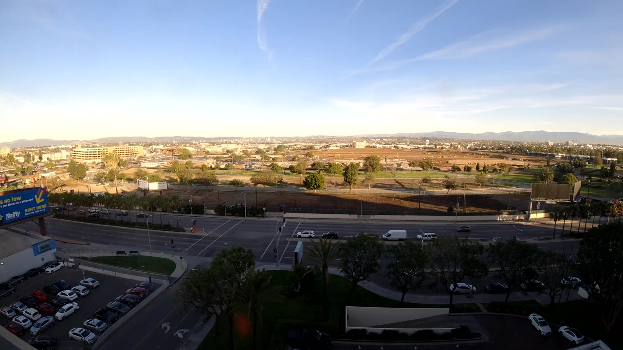 Elevated Los Angeles view at 0:00 with parked cars in the foreground, moving traffic on a multilane road, low buildings beyond, distant hills, and no visible people.