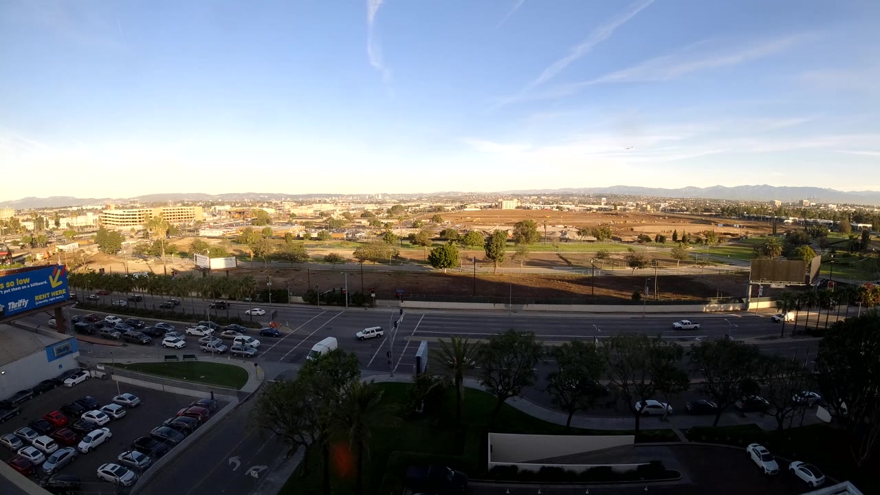At 0:02 the elevated Los Angeles frame shows a busier stretch of road with clustered cars, shaded trees in the foreground, commercial buildings, distant hills, and no visible people.