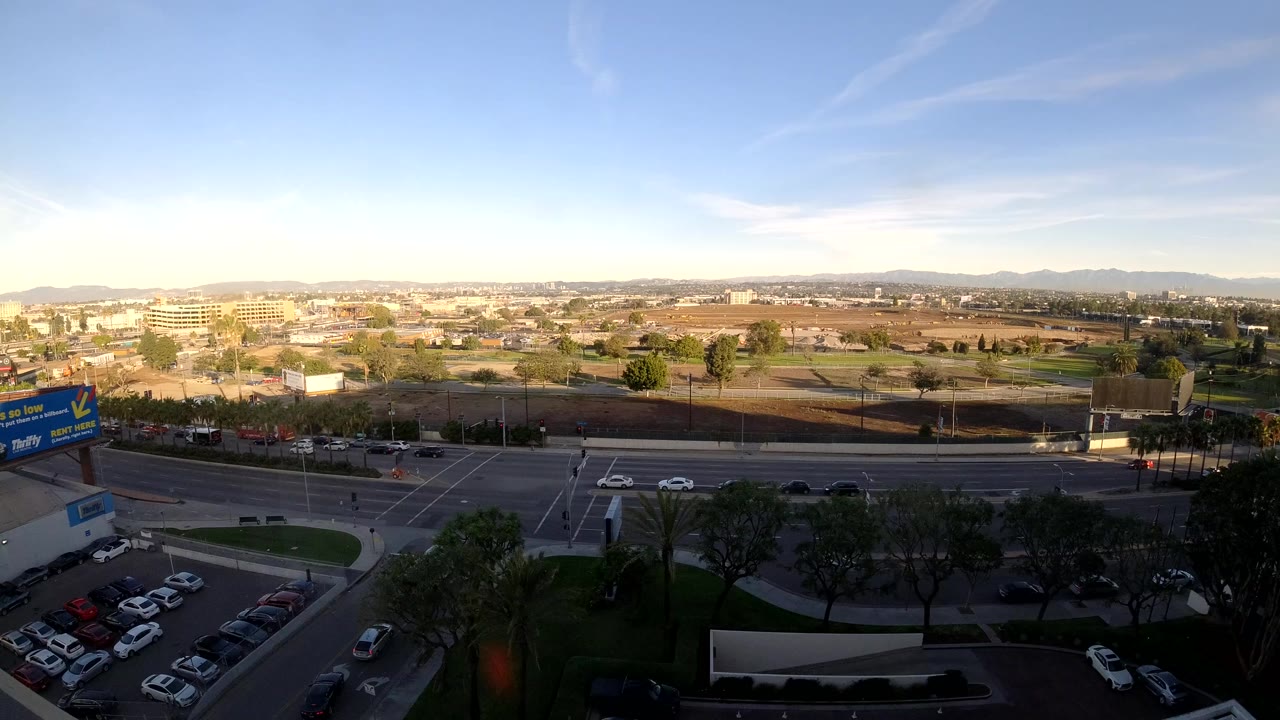 Los Angeles aerial-style frame with cars moving through the broad road and intersection, shaded trees in the foreground, low urban buildings in the distance, and no visible people.