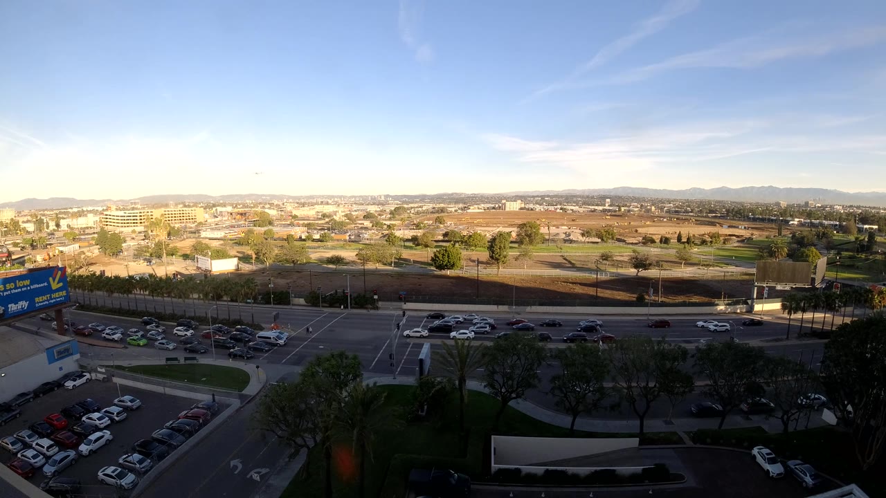 Elevated Los Angeles traffic view with several cars spread across the road, a shaded foreground of trees and parking, sunlit low buildings, distant hills, and no visible people.