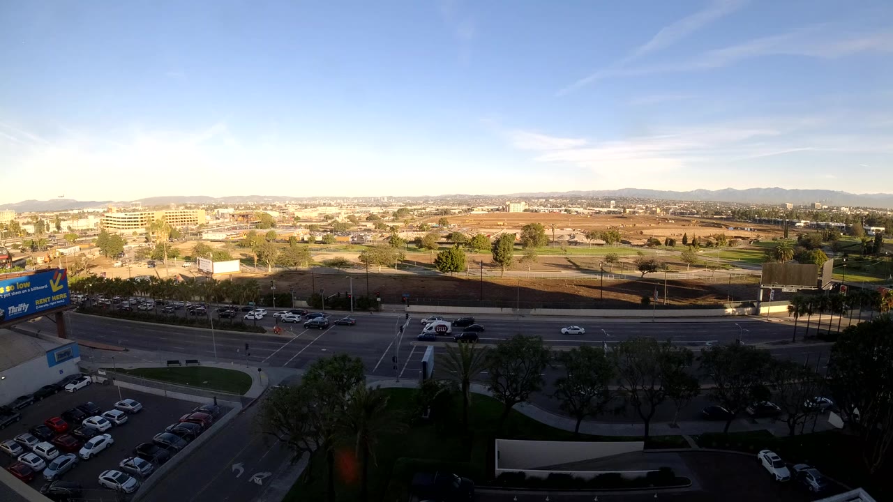 Start of a Los Angeles elevated traffic view with cars and trucks on a wide road, a parking lot in the foreground, distant hills on the horizon, and no visible people.