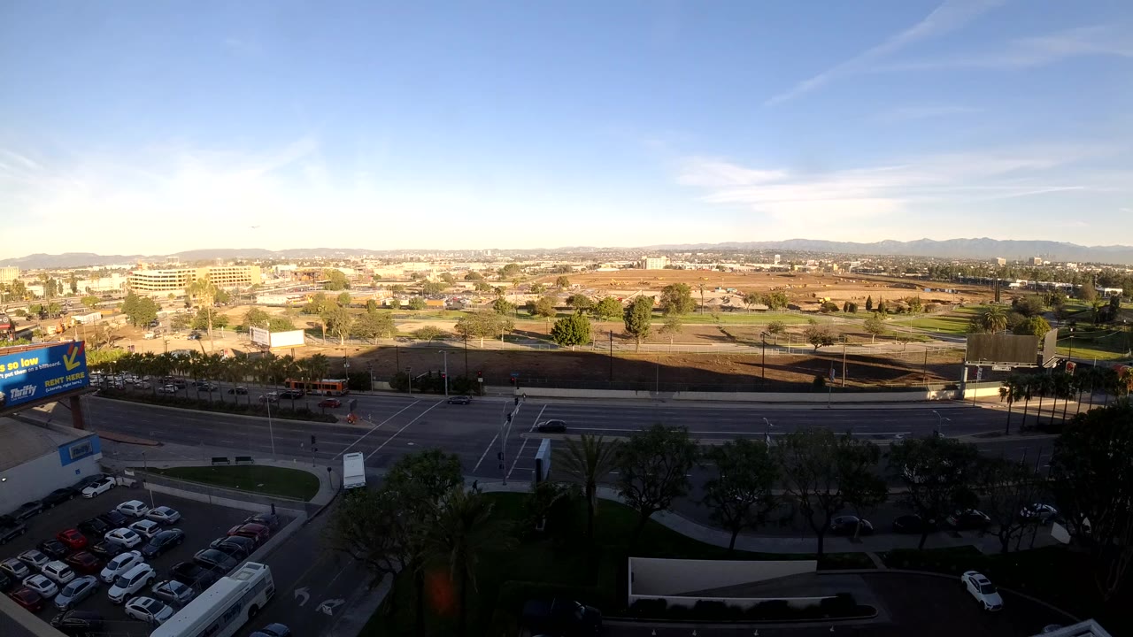 Elevated daylight view over Los Angeles traffic with a shaded parking lot in the foreground, a blue roadside billboard at left, distant low buildings, hills, and no visible people.