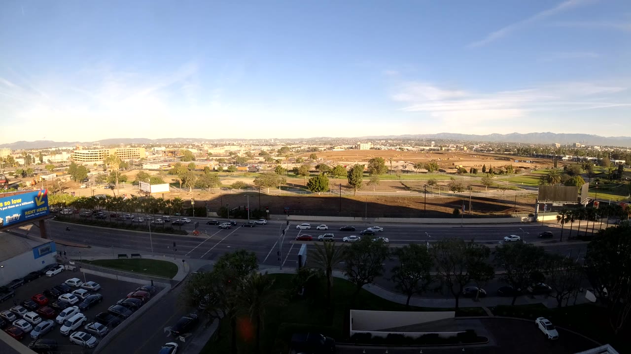 Wide elevated view of Los Angeles road traffic with a parking lot and trees in the foreground, distant hills under blue sky, and no visible people.