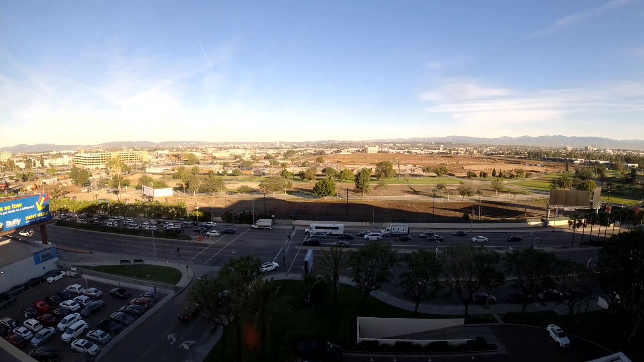 Start frame of Los Angeles traffic from an elevated view, showing cars on a multilane road, a parking lot in shadow, billboards, low buildings, distant mountains, and no visible people.