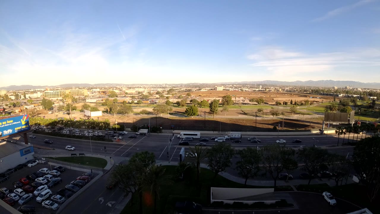 Los Angeles road traffic continues beneath bright late-day light, with cars in the midground, dark trees and parking below, billboards at left, mountains on the horizon, and no visible people.