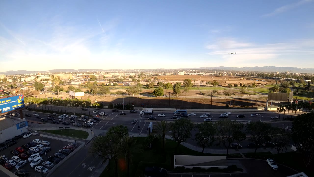 Mid-clip elevated view in Los Angeles with cars spread across the road, a visible parking lot, roadside billboards, low commercial buildings, a pale mountain ridge, and no visible people.