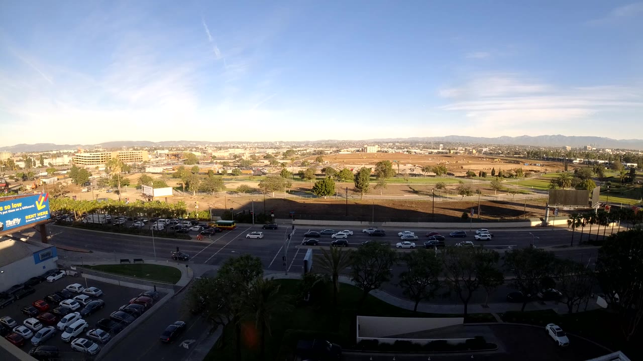 Wide daytime aerial view in Los Angeles showing an intersection, parking lot, commercial buildings, trees, distant hills, blue sky, and no visible people.
