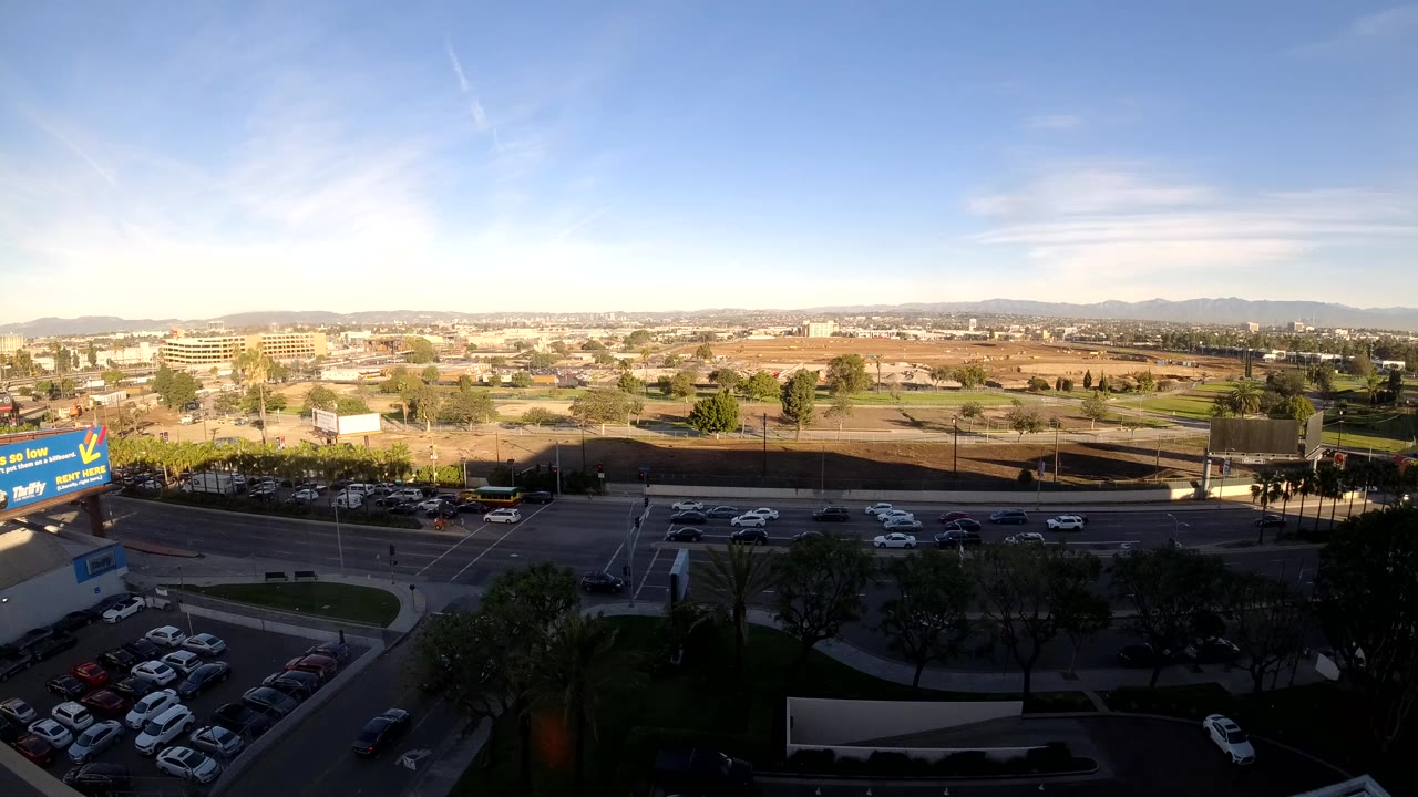 Elevated Los Angeles view with cars on a multilane road, rows of parked vehicles, low buildings, roadside signage, trees, distant hills, and no visible people.