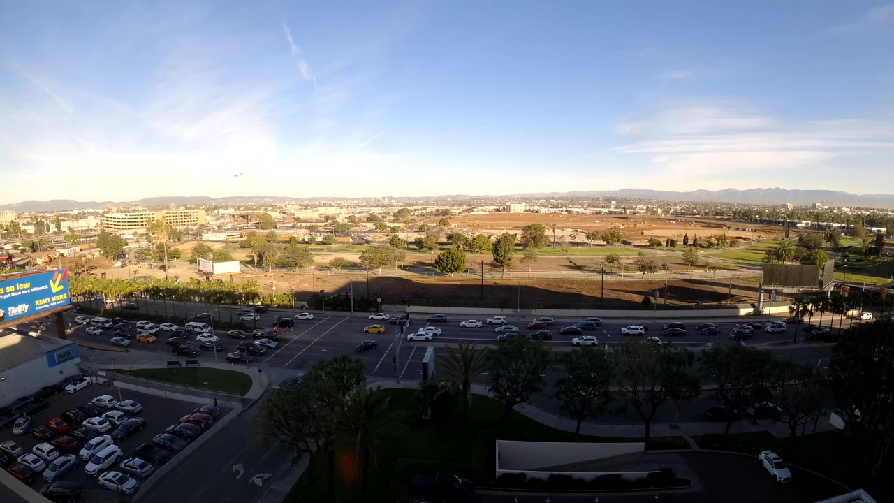Los Angeles aerial frame showing steady roadway traffic, a parking lot edge, shaded trees, commercial structures, distant hills, bright sky, and no visible people.